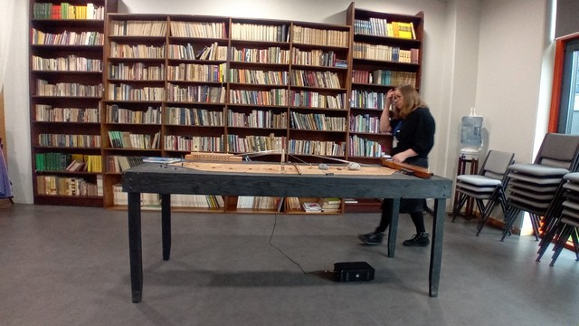 Liels grāmatu skapis pilns, priekšpusē liels koka galds.