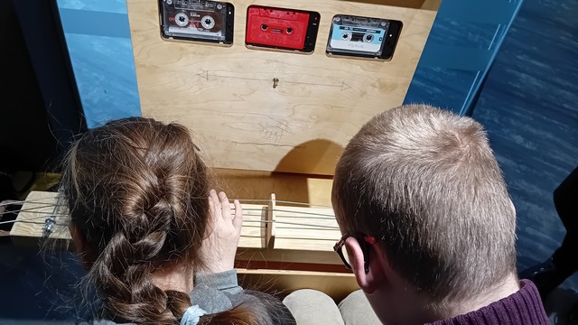 Zēns un meitene spēlē uz stīgām, pirkšā siena ar padziļinājumā esošām trim magnetafona kasetēm.
