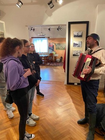 Skolēni stāv pretī vīrietim, kas ar ķiveri galvā spēlē akardeonu.