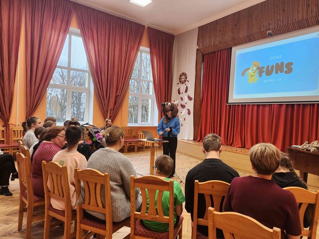 Skolēni skatās un klausās, vadītāja stāv pie datora stāsta.