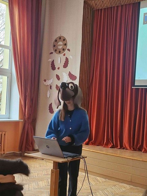 Pasākuma vadītāja pie datora ar suņa Fuņa masku.