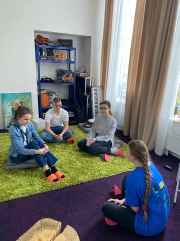 Skolēni aplī sasēdušies uz paklāja.