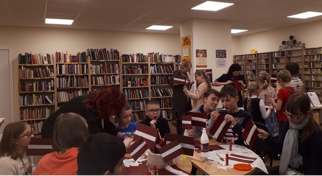 Bērni bibliotēkā pie galdiem veidot karodziņus. Skolotājas un bibblitekāres palīdz.