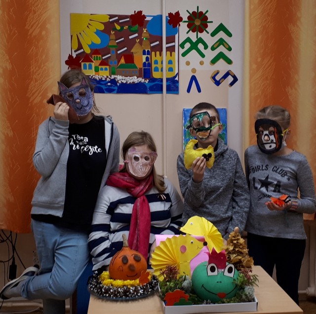 4 skolēni maskās sēž pie galda ar ķirbjiem un citiem izstādes eksponātiem