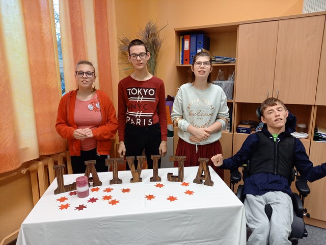 Četri skolēni pie galda ar no koka burtiem salikta uzraksta "Latvija". Uz galda svece un izgrieztas papīra zvaigznes.