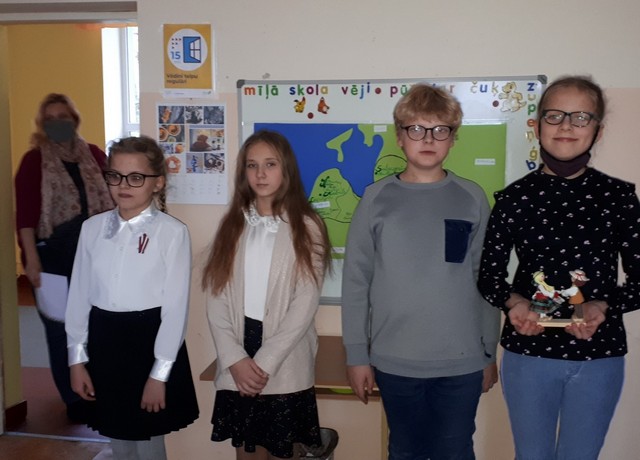 Četri 4.klases skolēni savā klasē pie Latvijas kartes. Skolotāja Vineta ienāk pa durvīm.