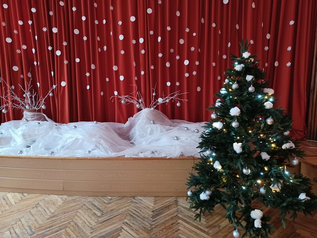 Skolas zālē - eglīte ar vates pikučiem . Uz skatuves audums izveidots kā sniegs. Aizkari arī ar baltām papīra sniegpārslām.