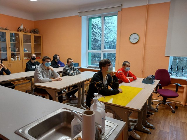 Skolēni Bioloģijas klasē sēž solos un klausās - skats no durvīm