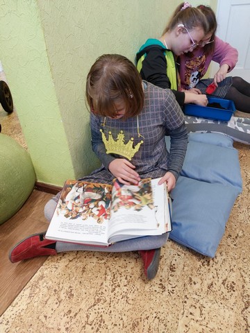 Priekšplānā meitene lasa grāmatu, tālāk divas meitenes darbojas ar zemi.