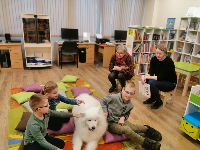 Bibliotēkā trīs skolēni glauda baltu suni. Bibliotēkas darbinieces sēž blakus ar grāmatām rokās.