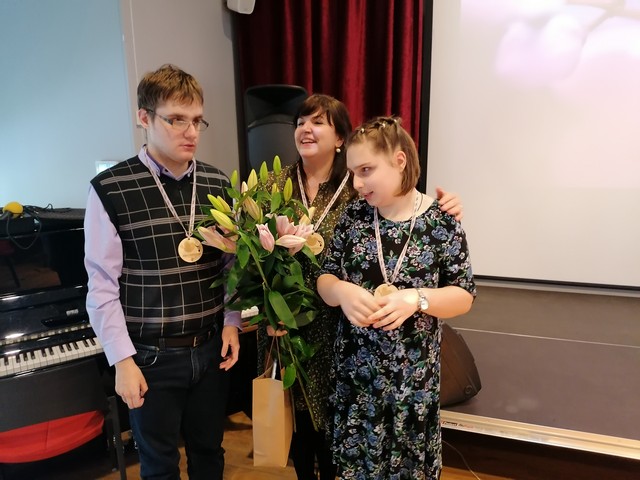 Divi mūsu skolas skolēni ar skolotāju Ivetu pie skatuves ar ziediem un dalībnieku medaļām.