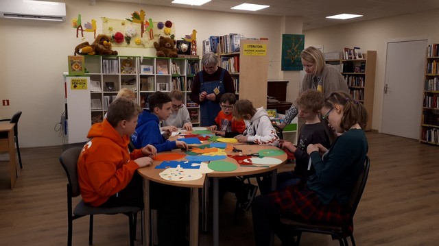 Divas bibliotēkas darbinieces palīdz bērniem, kas sēž pie apaļa galda, darboties ar krāsainām lapām.
