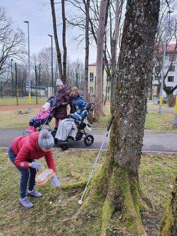 Skolotāja ar trim skolēniem pastaigājas, bet viena skolniece lasa olas, kas noslēptas pie liela koka.