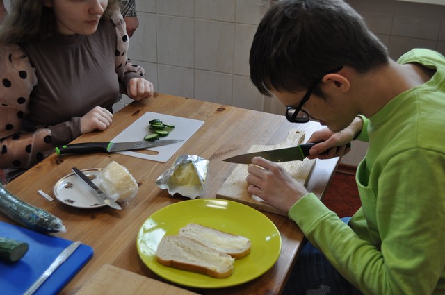 Divi skolēni ēdnīcā pie galda griež ar nažiem maizi un gurķus, smērē sviestu.