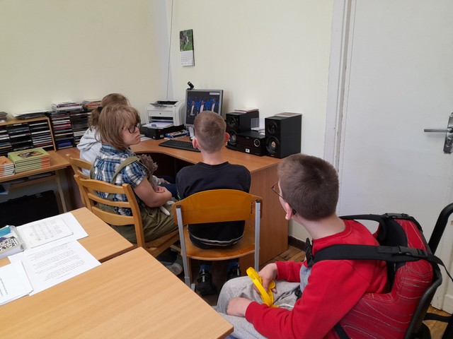 Trīs skolēni sēž pie datora un skatās video ar himnu, viens zēns tālāk ratiņkrēslā ar skatās un klausās.