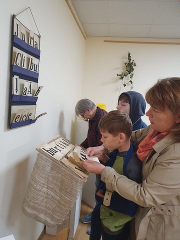 Skolotājas Inese un Gundega kopā ar skolēniem apskata lapiņas ar lieliem burtiem.