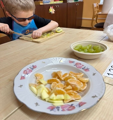 Zēns pie galad griež augļus. Divi šķīvji ar vīnogām un mandarīniem.