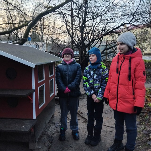 Trīs skolēni stāv pie kaķu mājas.