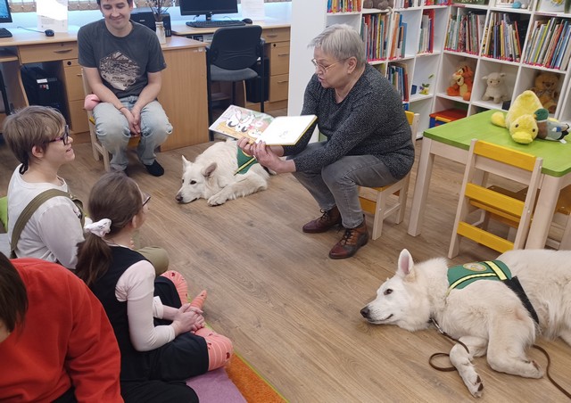 Bibliotekāre Gundega rāda skolēniem atvērtu grāmatu, abās pusēs viņai guļ balti pūkaini suņi.