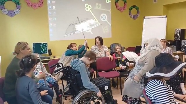 Visi sēž aplī ap vecmāmiņu. Skolotāja Agnese atvērusi lietussargu.