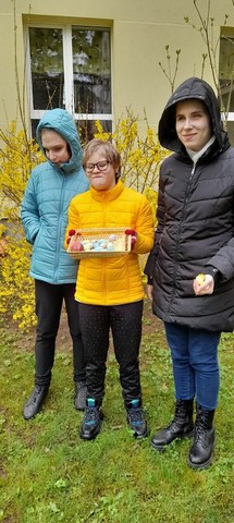 Trīs meitenes stāv skolas pagalmā, vidējā tur paplāti ar krāsotām olām.