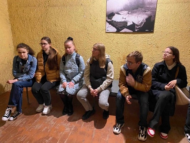 Skolēni sēž uz gara sola pie grubuļainas sienas.