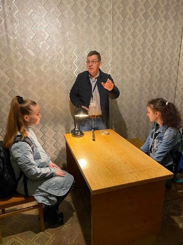 Divas meitenes sēz pie galda, gids stāsta.