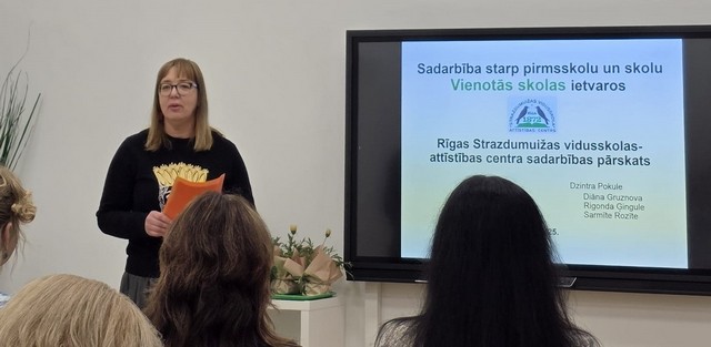 Dzintra Pokule pie interaktīvā ekrāna, uz krua ir skolas nosaukums "Rīgas Strazdumuižas vidusskolas - attīstības centra sadarbības pārskats.