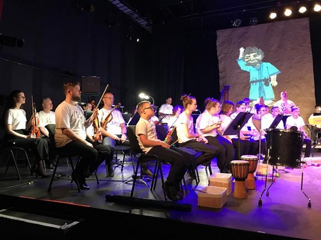 Izrādes mūziķi uz sēž skatuves, tālāk erāns ar zīmē'jumu.