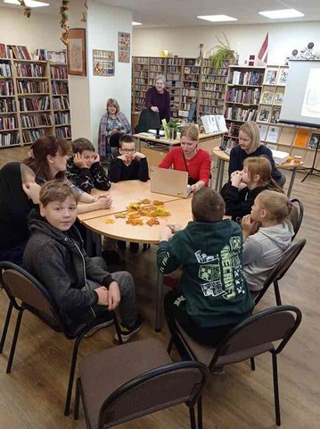 Skolēni, skolotāja un divas bibliotēkas darbinieces sēž pie apaļa galda.
