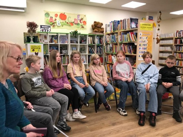 Skolēni pusaplī sēž uz krēsliem bibliotēkā.