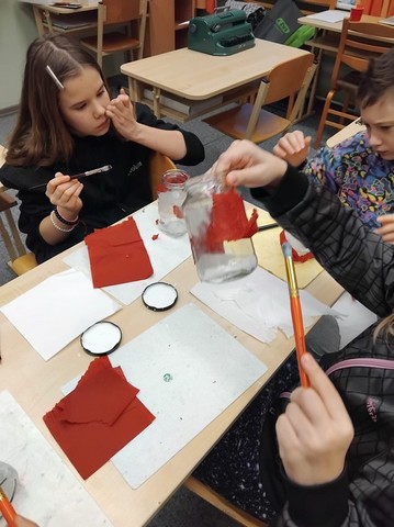 Skolēni pie galda krāso ar sarkano un balto krāsu.