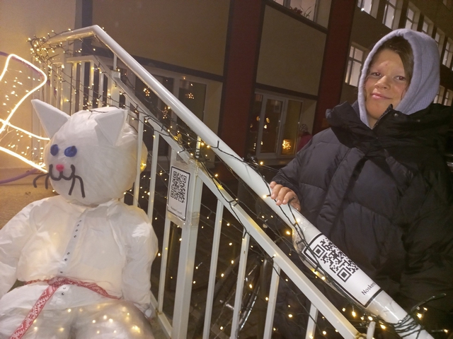 Meitene stāv uz trepēm ar gaismiņām, prie kurām ir arī uztaisīts balts liels kaķis.