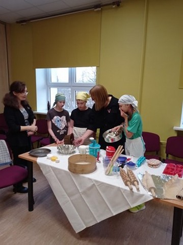 Skolotājas Dzintra un Elita pie galda 3 skolēniem rāda kūkas gatavošanas iekārtas.