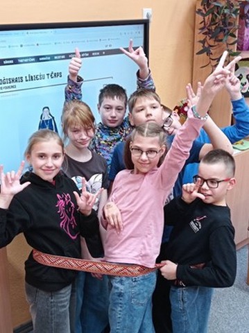 Skolēnu grupiņa klaē pie interaktīvā ekrāna, apvijušies ar sarkanu jostu.