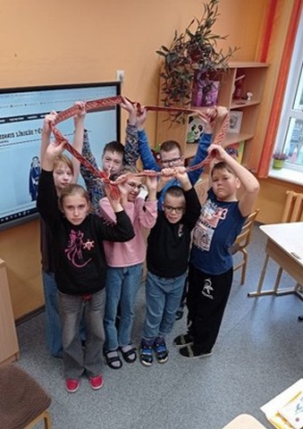 Skolēnu grupa klasē tur virs galvas sarkanu jostu.