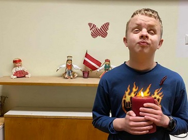 Alberts stāv klasē pie sienas, aiz muguras Latvijas karodziņš, rokās tur lielu aizdegtu sveci.