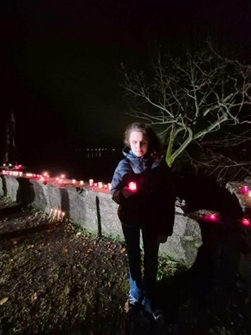 Lelde stāv pie mūra ar svecītēm un pati tur rokā aizdegtu svecīti.