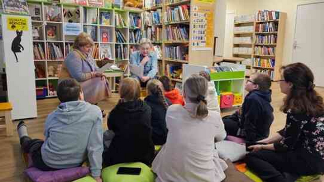 Divas Bibliotēkas darbinieces lasa bērniem un divām skolotājām, kas sasēdušās uz grīdas.