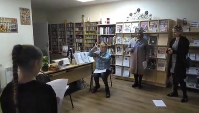 Bibliotēkas darbinieces stāsta, viena sēž, divas stāv.