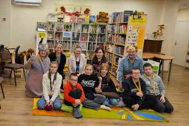 Visi pasākuma dalibnieki sēž uz grīdas bibliotēkā.