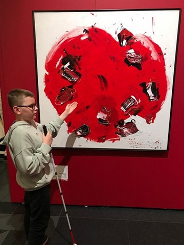 Zēns ar balto spieķi stāv pie lielas gleznas.