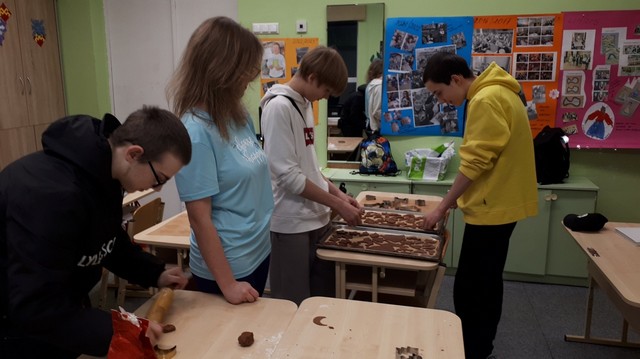 Divi zēni kārto pannās piparkūkas, viens zēns rullē mīklu, meitene stāv pa vidu un skatās.