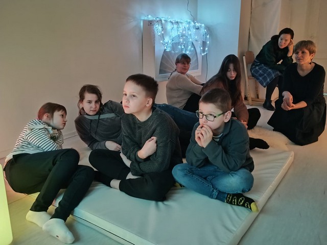 Pieci skolēni un trīs skolotājas sēž uz matrača, grīdas un klausās.