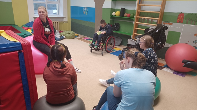 Skolotāja Agrita stāsta skolēniem, kas apsēdušies ap viņu.