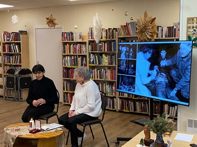 Velta Puriņa un bibliotekāre Gundega sēž pie apaļa galdiņa, aiz viņām ekrāns, kas rāda fotogrāfijas.