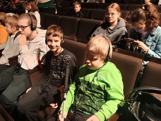 Skolēni sēž teātra zālē ar austiņām.