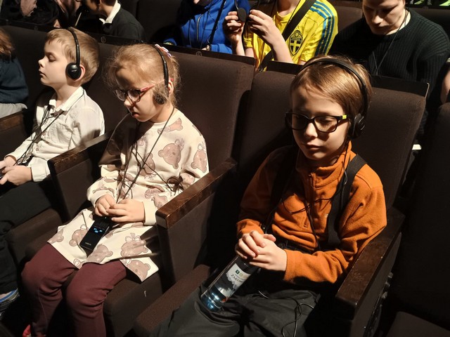 Divi zēni un viena meitene sēž teātra zālē ar austiņām.