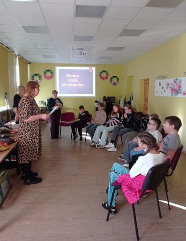 Skolotāja Dzintra konferenču zālē lasa bērniem, kas sēž pusaplī apkārt.