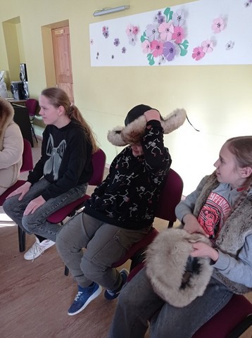 Skolēni sēž, viens liek galvā ausaini, otra tur rokās zvērādas cepuri.